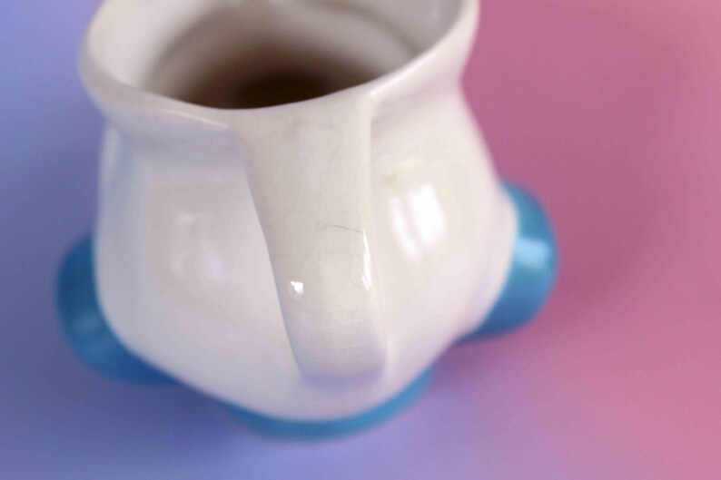 May include: A white ceramic pitcher featuring a glossy finish with visible fine crackle patterns throughout the glaze. The pitcher has a curved handle and is supported by three rounded, bright blue feet positioned at the base of the vessel.
