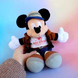 May include: A Mickey Mouse plush toy wearing a brown jacket, blue jeans, and a tan hat with a blue brim. The toy has black ears, white gloves, and brown and gray shoes. The shirt says "The Walt Disney Studios".