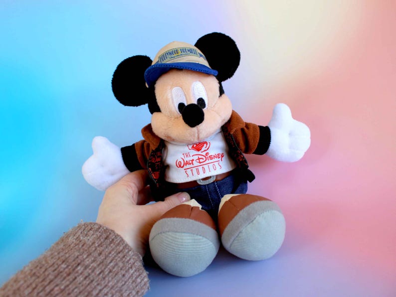 May include: A Mickey Mouse plush toy wearing a brown jacket, blue jeans, and a tan hat with a blue brim. The toy has black ears, white gloves, and brown and gray shoes. The shirt says "The Walt Disney Studios".