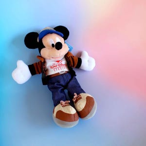 May include: A plush Mickey Mouse doll wearing a brown jacket, blue jeans, and a blue cap. The doll has white gloves and brown and white shoes. The shirt says "Walt Disney Studios". The background is a gradient of blue, pink, and orange.