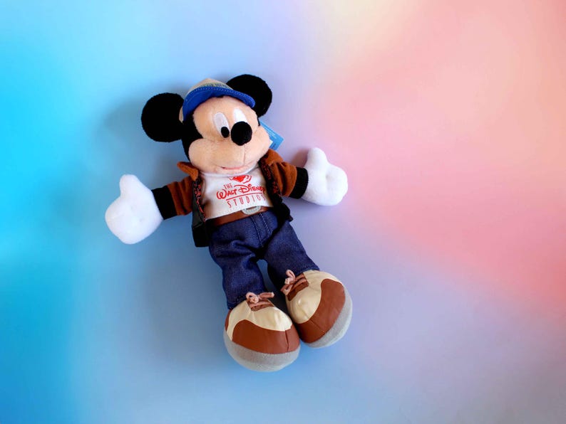 May include: A plush Mickey Mouse doll wearing a brown jacket, blue jeans, and a blue cap. The doll has white gloves and brown and white shoes. The shirt says "Walt Disney Studios". The background is a gradient of blue, pink, and orange.