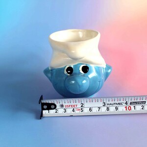 May include: A blue and white ceramic mug shaped like a Smurf character head, featuring a white hat and large eyes. A metal measuring tape is positioned in front of the mug, showing a width of approximately 3.5 inches at the widest point.