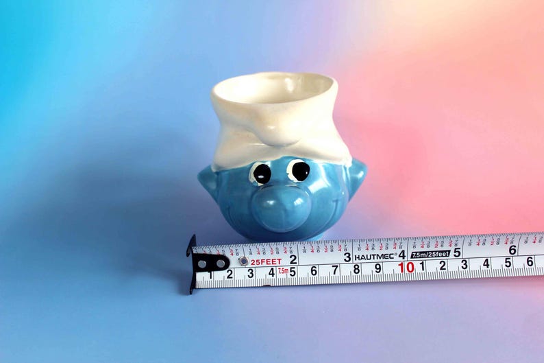 May include: A blue and white ceramic mug shaped like a Smurf character head, featuring a white hat and large eyes. A metal measuring tape is positioned in front of the mug, showing a width of approximately 3.5 inches at the widest point.