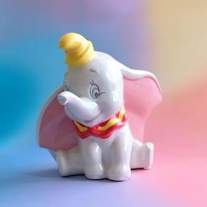May include: A white ceramic Dumbo the elephant figurine with a yellow hat, pink ears, and a red and yellow collar. The figurine is set against a soft, colourful background, evoking a whimsical and nostalgic feel.