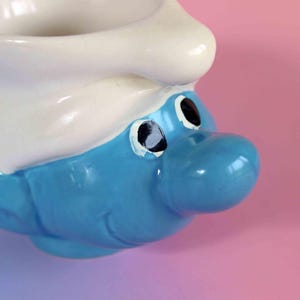 May include: A ceramic mug shaped like a Smurf character head, featuring a bright blue face with a large nose, black eyes, and a white hat that forms the rim of the cup. The mug has a smooth, glossy finish and a handle on the side for holding.