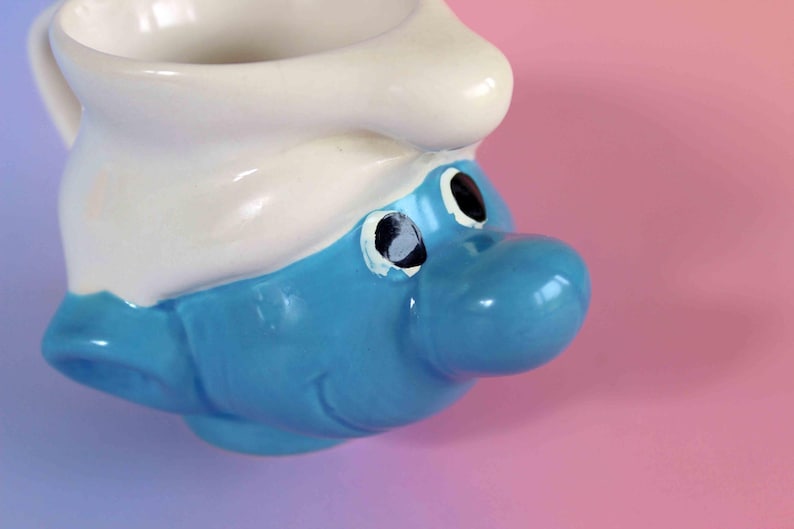 May include: A ceramic mug shaped like a Smurf character head, featuring a bright blue face with a large nose, black eyes, and a white hat that forms the rim of the cup. The mug has a smooth, glossy finish and a handle on the side for holding.