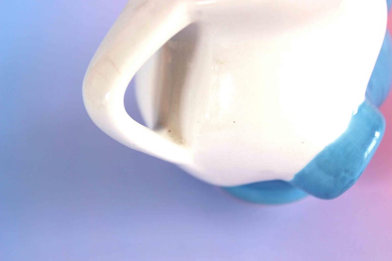 May include: A white ceramic pitcher featuring a curved handle and a smooth, glossy finish. The base of the pitcher is accented with a vibrant blue glaze, creating a distinct contrast against the white body. The design is simple and functional.