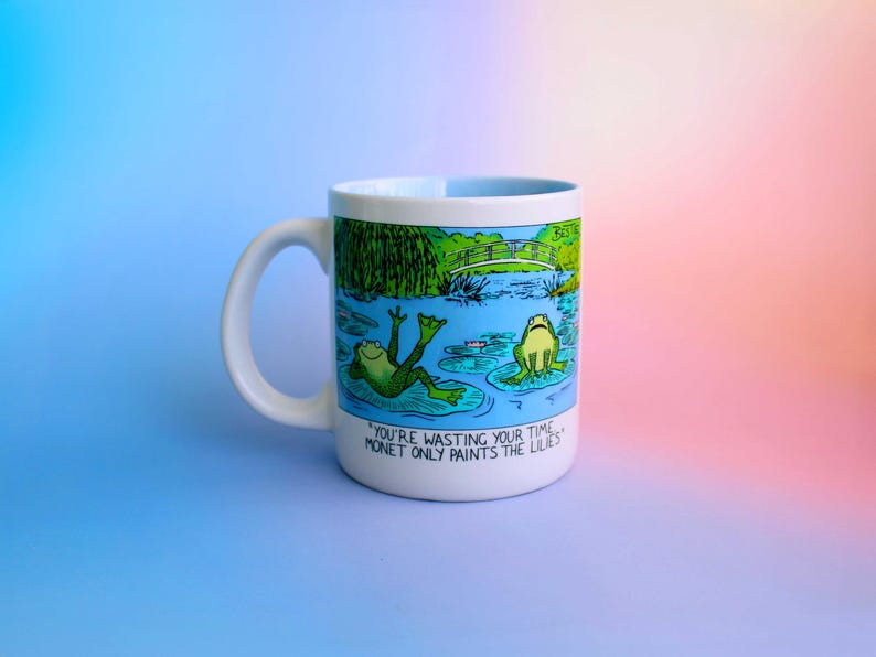 K&ouml;nnte beinhalten: Wei&szlig;e Keramik-Tasse mit einer Cartoon-Illustration von zwei Fr&ouml;schen in einem Teich mit Seerosen und einer Br&uuml;cke. Auf der Tasse steht der Text "YOU'RE WASTING YOUR TIME. MONET ONLY PAINTS THE LILIES."
