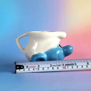 May include: A ceramic mug shaped like a Smurf character with a white hat and blue face, positioned next to a metal measuring tape showing a length of approximately 5.5 inches from the handle to the nose of the character.