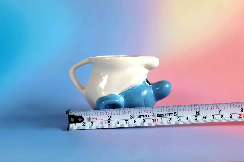 May include: A ceramic mug shaped like a Smurf character with a white hat and blue face, positioned next to a metal measuring tape showing a length of approximately 5.5 inches from the handle to the nose of the character.