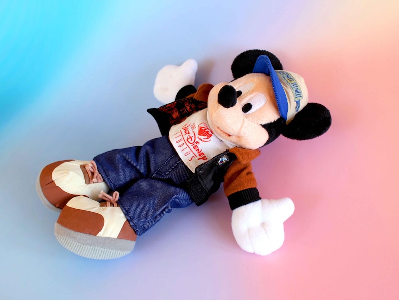 May include: A plush Mickey Mouse doll wearing a blue and white cap, a brown and black jacket, blue jeans, and brown and white shoes. The doll has a white shirt with "The Disney Studios" printed on it. The background is a gradient of pink and blue.