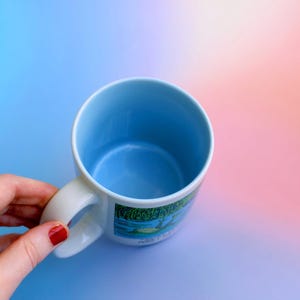 K&ouml;nnte beinhalten: Eine wei&szlig;e Keramiktasse mit hellblauem Innenraum und Henkel. Die Tasse hat ein farbenfrohes Landschaftsdesign auf der Au&szlig;enseite. Die Tasse wird von einer Hand mit rotem Nagellack vor einem Farbverlaufshintergrund gehalten.
