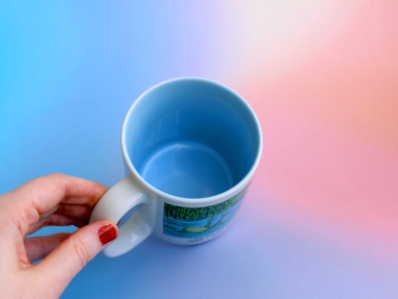 K&ouml;nnte beinhalten: Eine wei&szlig;e Keramiktasse mit hellblauem Innenraum und Henkel. Die Tasse hat ein farbenfrohes Landschaftsdesign auf der Au&szlig;enseite. Die Tasse wird von einer Hand mit rotem Nagellack vor einem Farbverlaufshintergrund gehalten.