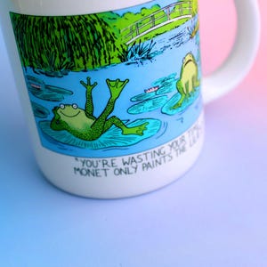 K&ouml;nnte beinhalten: Wei&szlig;e Keramiktasse mit einer Cartoon-Illustration von Fr&ouml;schen auf Seerosenbl&auml;ttern in einem Teich, mit einer Br&uuml;cke und Gr&uuml;n in der Hintergrund. Die Tasse hat den Text "YOU'RE WASTING YOUR TIME MONET ONLY PAINTS THE LILIES."