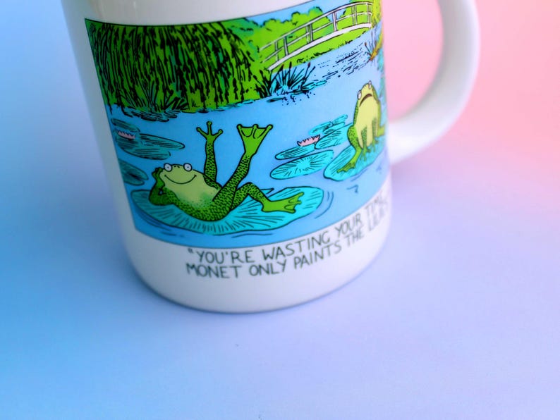 K&ouml;nnte beinhalten: Wei&szlig;e Keramiktasse mit einer Cartoon-Illustration von Fr&ouml;schen auf Seerosenbl&auml;ttern in einem Teich, mit einer Br&uuml;cke und Gr&uuml;n in der Hintergrund. Die Tasse hat den Text "YOU'RE WASTING YOUR TIME MONET ONLY PAINTS THE LILIES."