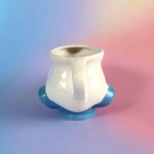 May include: A ceramic mug featuring a white upper body and a bright blue base with two rounded blue protrusions on the sides. The mug has a single handle on the right side and a wide, open top. The surface is smooth and glossy with a reflective finish.