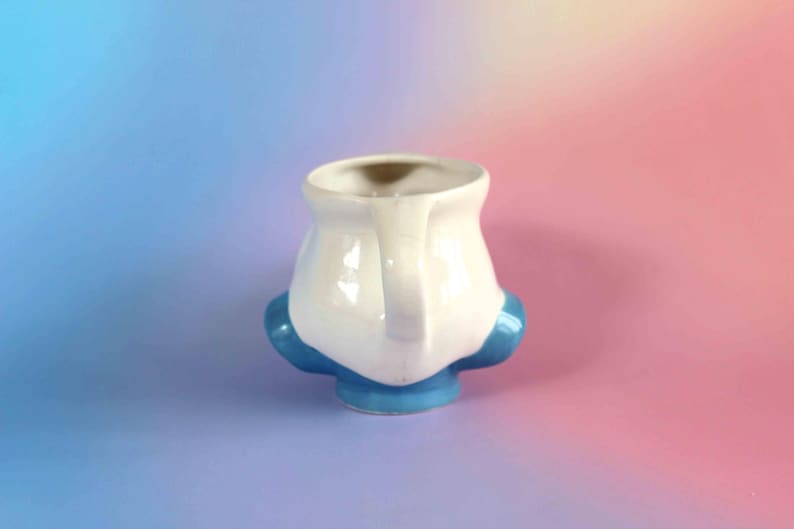 May include: A ceramic mug featuring a white upper body and a bright blue base with two rounded blue protrusions on the sides. The mug has a single handle on the right side and a wide, open top. The surface is smooth and glossy with a reflective finish.
