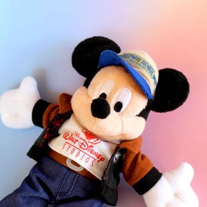 May include: A plush Mickey Mouse doll wearing a brown jacket, blue jeans, and a blue and tan cap. The doll has black ears, a black nose, and white gloves. The shirt says "The Walt Disney Studios".
