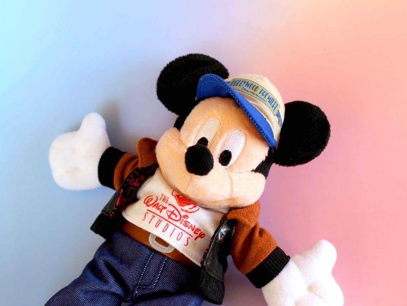 May include: A plush Mickey Mouse doll wearing a brown jacket, blue jeans, and a blue and tan cap. The doll has black ears, a black nose, and white gloves. The shirt says "The Walt Disney Studios".