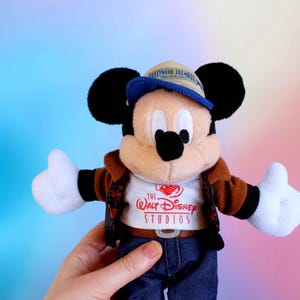 May include: A Mickey Mouse plush toy wearing a brown jacket, blue jeans, and a blue and tan cap. The white t-shirt features "The Walt Disney Studios" logo. The plush has black ears, a black nose, and white gloves.