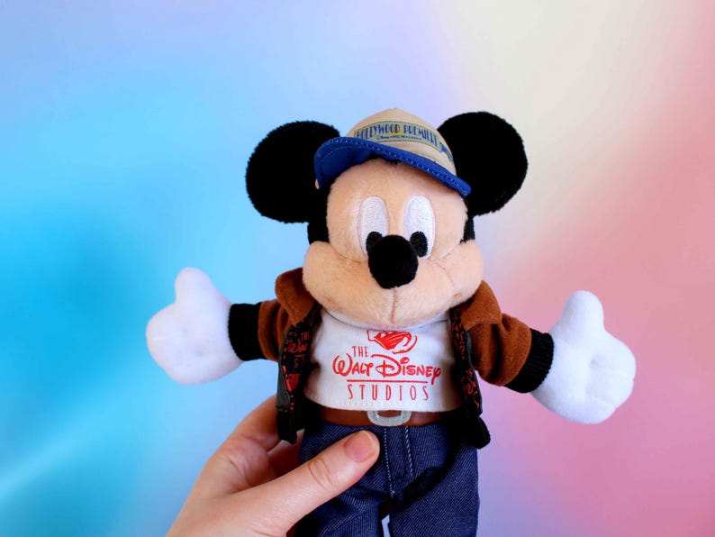May include: A Mickey Mouse plush toy wearing a brown jacket, blue jeans, and a blue and tan cap. The white t-shirt features "The Walt Disney Studios" logo. The plush has black ears, a black nose, and white gloves.
