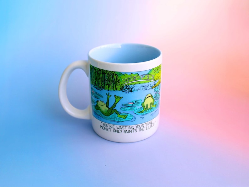 May include: White ceramic mug with a light blue interior and a frog pond illustration. The design features two green frogs on lily pads, with the text "You're wasting your time. Monet only paints the lilies."