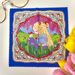 May include: A square microfibre cleaning cloth with a blue border and pinked edges. The central illustration features a blonde elf with braided hair in a blue cloak, surrounded by green foliage, red mushrooms, and purple wisteria flowers.