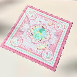 May include: A square microfiber cleaning cloth with pink scalloped edges, featuring a central illustration of a person in a green sweater and yellow skirt surrounded by tulips, small animals, and circular floral motifs on a white background.
