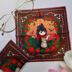 May include: A square microfiber cleaning cloth with a scalloped edge featuring a black and white cat illustration wearing a red scarf and apron, surrounded by a wreath of red flowers and butterflies on an orange and dark brown patterned background.