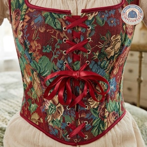 May include: A corset featuring a deep red tapestry fabric with a multi-colored floral pattern in shades of green, blue, and gold. The front includes silver-toned metal grommets with a red satin ribbon lace-up closure tied in a bow at the center.