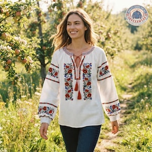 May include: A long-sleeved white blouse featuring a V-neckline with red tassel ties and intricate floral embroidery in red, blue, and green along the chest and sleeves. The garment has a relaxed fit and is paired with dark blue denim jeans.
