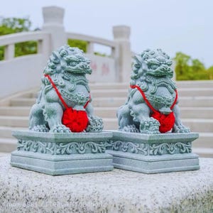 May include: Two grey stone lion statues with red details. Each lion sits on a decorative base and wears a red collar with a red ball. The statues feature detailed carvings and are positioned outdoors, possibly in a garden.