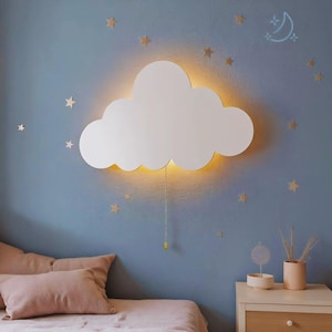 Peut inclure: Une applique murale blanche en forme de nuage avec une lueur chaude, accrochée à un mur bleu. La lumière a une chaîne à tirer et est entourée d'étoiles dorées. Une table de chevet avec une lampe et un lit avec des oreillers roses sont au premier plan.