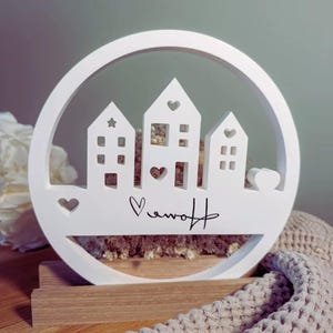 Könnte beinhalten: Weißes Holzdekor mit drei ausgeschnittenen Häusern in einem kreisförmigen Rahmen. Die Häuser haben Herz- und Sternformen, mit dem Wort "Home" in Schrift. Es steht auf einem Holzsockel.