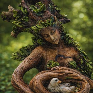 May include: A brown, tree-like sculpture of a human figure with closed eyes, adorned with green leaves. The figure holds a hollowed-out wooden section containing a small, light-colored bird with a yellow beak resting inside a nest-like structure.