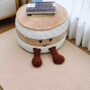 May include: A round, plush ottoman in a whimsical design, resembling a layered dessert. The top is tan, with white and tan layers, and brown legs. A small stack of books rests on top. The ottoman has a smiling face.