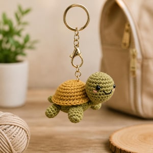 May include: A crochet turtle keychain featuring a green head and limbs with a mustard yellow shell. The turtle is attached to a gold-toned metal split ring and lobster clasp. The turtle has small black bead eyes and embroidered pink cheeks.