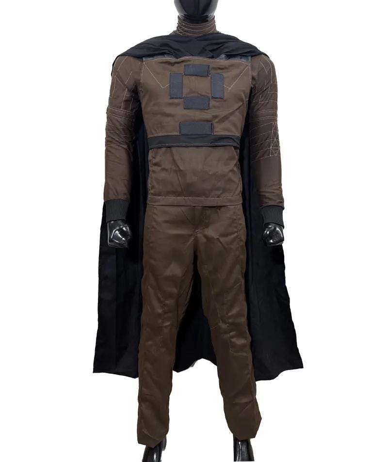 May include: A brown costume with a black cape. The costume includes a long-sleeved jacket with square patches, matching pants, and a black cowl. The cape is attached at the shoulders. The outfit is displayed on a mannequin.