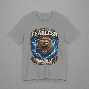 May include: Gray t-shirt featuring a bear graphic with the text "FEARLESS" above and "CONQUER ALL" below. The bear is surrounded by blue lightning bolts, with an American flag in the background. The shirt also includes the text "BUILT TO ENDURE" and "NEVER BACK DOWN".
