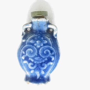 May include: A small, blue ceramic bottle with a decorative floral design. The bottle has a cylindrical neck and a small, green stopper. The body of the bottle is a deep blue with white accents.