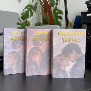 Op de afbeelding: Drie gebonden boeken met de titels Onyx Storm, Iron Flame en Fourth Wing. Elk boek heeft een lichte kaft met een romantische illustratie van twee personen en goudkleurige letters voor de titels, opgesteld op een zwarte tafel.