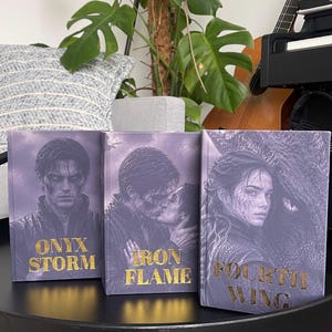 Peut inclure: Trois livres à couverture rigide aux tons violets avec des illustrations de personnages et du texte en feuille d'or. Les titres sont Onyx Storm, Iron Flame et Fourth Wing. Chaque couverture présente des illustrations détaillées de personnages.