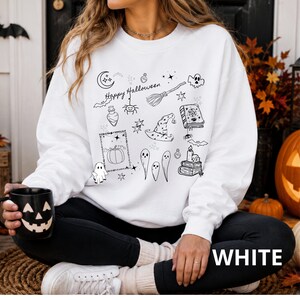 May include: A white long-sleeve sweatshirt featuring a black line art graphic print of Halloween-themed icons including a crescent moon, a broomstick, a witch hat, a spellbook, ghosts, a pumpkin, and the text "Happy Halloween" across the center.