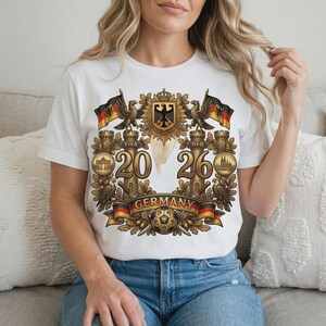 May include: White t-shirt featuring a detailed graphic with the text "2026 GERMANY". The design includes German flags, eagles, a soccer ball, and the German coat of arms. The shirt is made of a soft material and is suitable for casual wear.