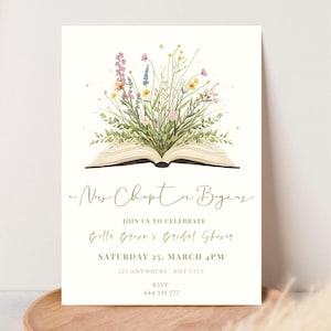 Puede incluir: Invitación a una despedida de soltera con el texto «Nuevo capítulo comienza». La invitación presenta un libro abierto con una ilustración de acuarela de flores silvestres. Detalles: fecha, hora, lugar e información RSVP.