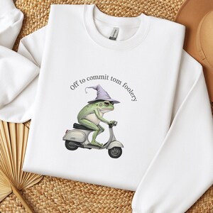 May include: White sweatshirt featuring a whimsical illustration of a green frog wearing a wizard hat and riding a silver scooter. The text "Off to commit tom foolery" is arched above the frog. The sweatshirt has a crew neck and long sleeves.