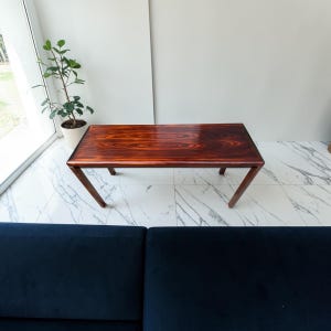 May include: A rectangular coffee table featuring a polished, dark reddish-brown wood grain surface with prominent, swirling patterns. The table is supported by four angled wooden legs that extend outward from the corners of the tabletop.