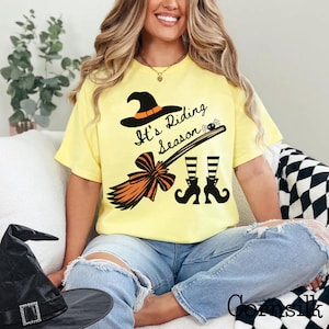 May include: A pale yellow short-sleeve t-shirt featuring a black graphic print of a witch hat, a broomstick, and striped stockings. The text "It's Riding Season" is printed in black script above the broomstick, accompanied by a small spider.