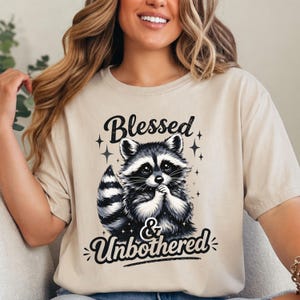 May include: Beige t-shirt with a black and white raccoon graphic. The text "Blessed & Unbothered" is above and below the raccoon. The shirt has a relaxed fit and short sleeves.