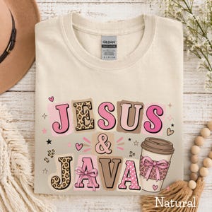 May include: Cream-colored t-shirt with the words "JESUS & JAVA" in pink, brown, and leopard print. A coffee cup with a leopard print bow is also featured. The shirt is made of heavy cotton.